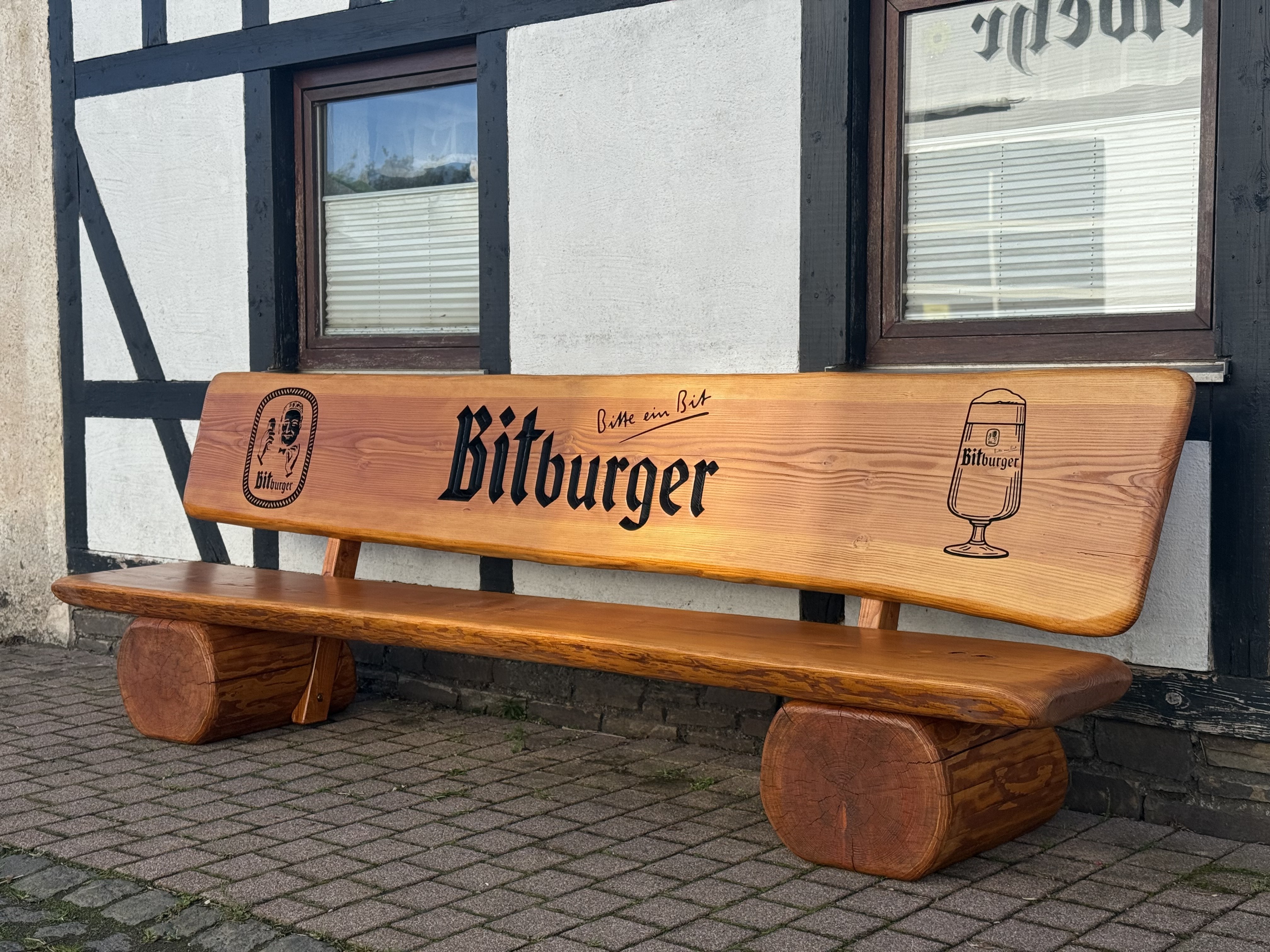 Rustikale Baumstammbank mit Bitburger Logo-Gravur vor Fachwerkhaus